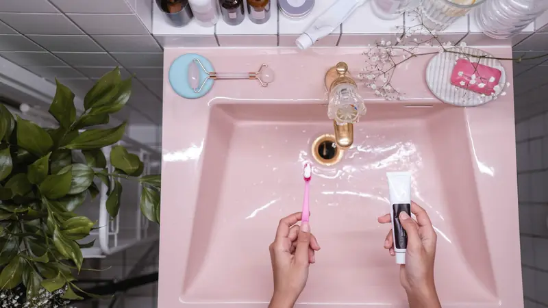 Mains tenant une brosse à dents garnie de dentifrice et un tube au-dessus d'un lavabo rose, vu de dessus