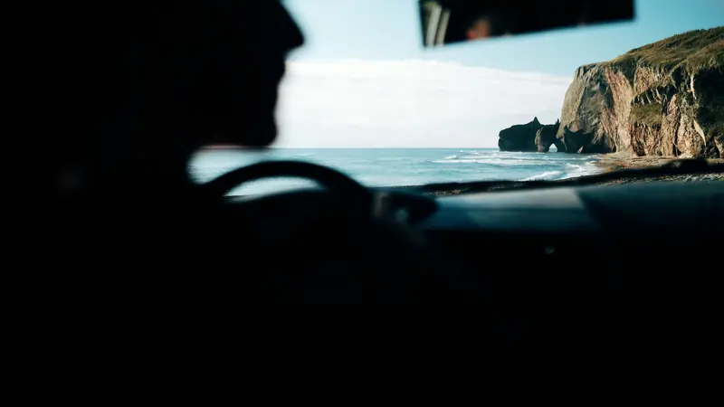 Conducteur en silhouette au volant face à une mer lumineuse et à une falaise, vu depuis l'habitacle