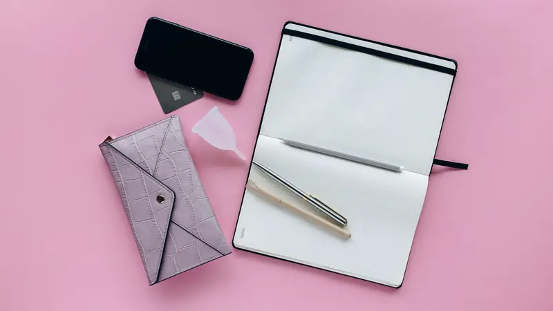Carnet quadrillé ouvert avec des stylos, un téléphone et une coupe menstruelle sur fond rose