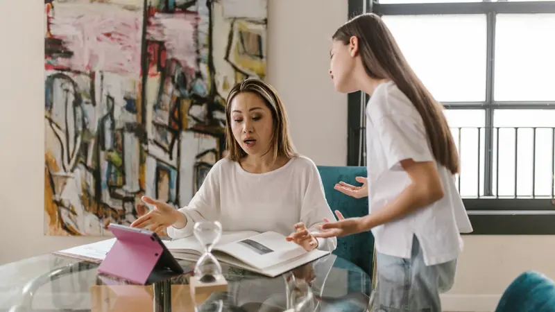 Une mère assise devant une tablette et un livre ouvert discute avec sa fille debout à côté de la table