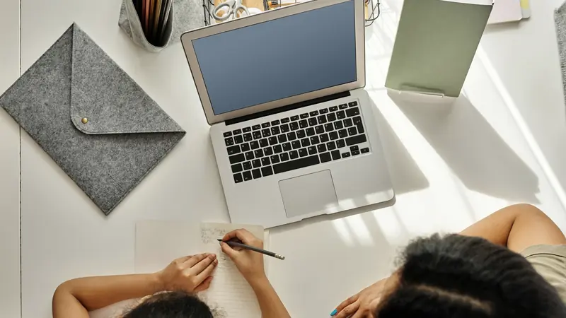 Vue de dessus d'un bureau blanc avec un ordinateur portable ouvert, un enfant qui écrit dans un cahier et un adulte assis à côté de lui