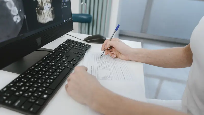 Mains remplissant un schéma dentaire sur papier devant un écran affichant une imagerie de la mâchoire