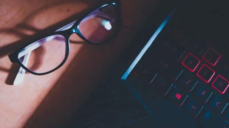 Lunettes de vue à monture noire posées sur un bureau en bois à côté du clavier rétroéclairé d'un ordinateur portable