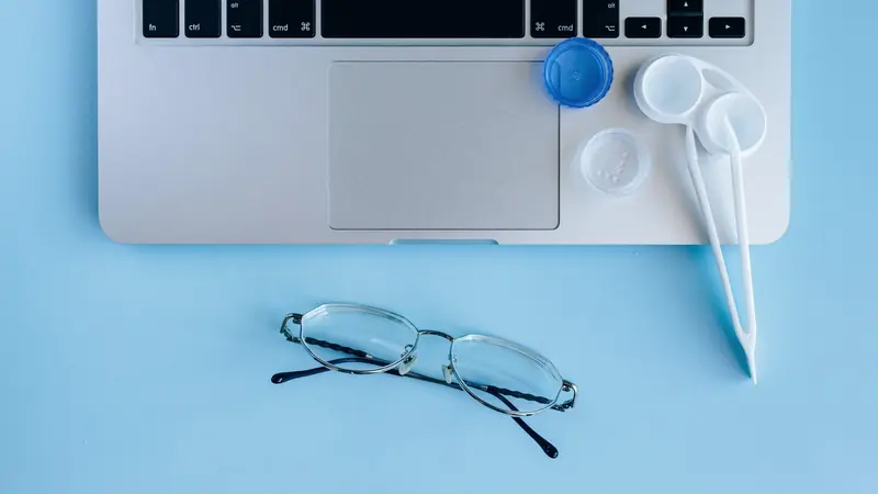 Lunettes à monture métallique posées devant un ordinateur portable, à côté d'un étui à lentilles, sur fond bleu