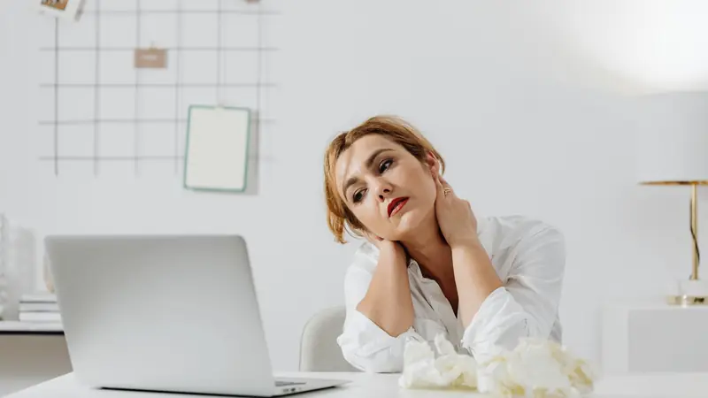 Femme fatiguée assise devant un ordinateur portable, la main sur la nuque, des mouchoirs froissés sur la table