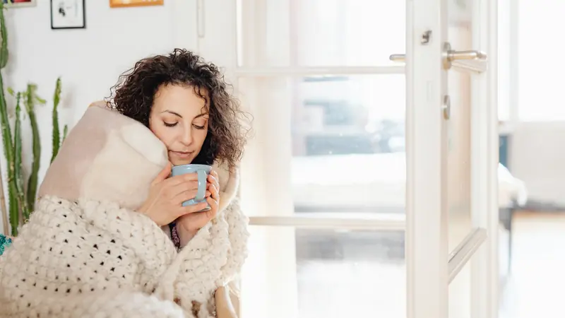 Femme enveloppée dans une couverture en laine qui tient une tasse chaude près d'une porte vitrée