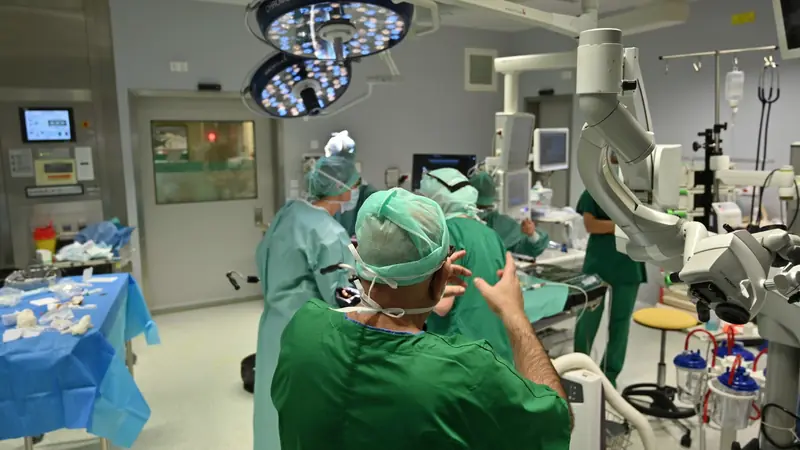 Équipe médicale en blouses vertes et charlottes autour d'une table d'intervention, sous les scialytiques d'une salle hospitalière équipée d'écrans
